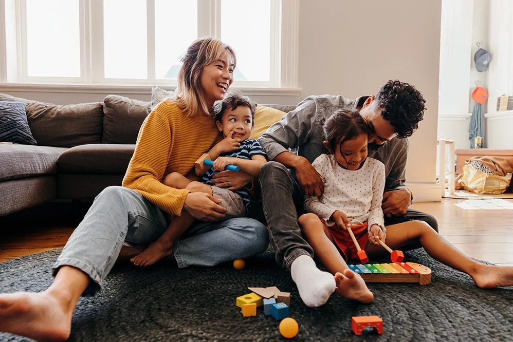 A family with medium-light skin tone sit on the living room floor, parents hugging their two children.